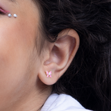 Tiny Jewels close-up of an ear with pink flower-shaped earrings and a visible earring in the background.