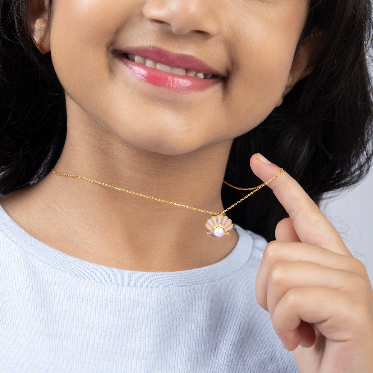 Tiny Jewels woman wearing a gold necklace with a shell pendant, pointing to it.