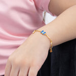Tiny Jewels gold bracelet with blue accents on a wrist against a pink and black background