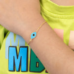 Tiny Jewels gold bracelet with a blue charm on a wrist, against a green shirt background.
