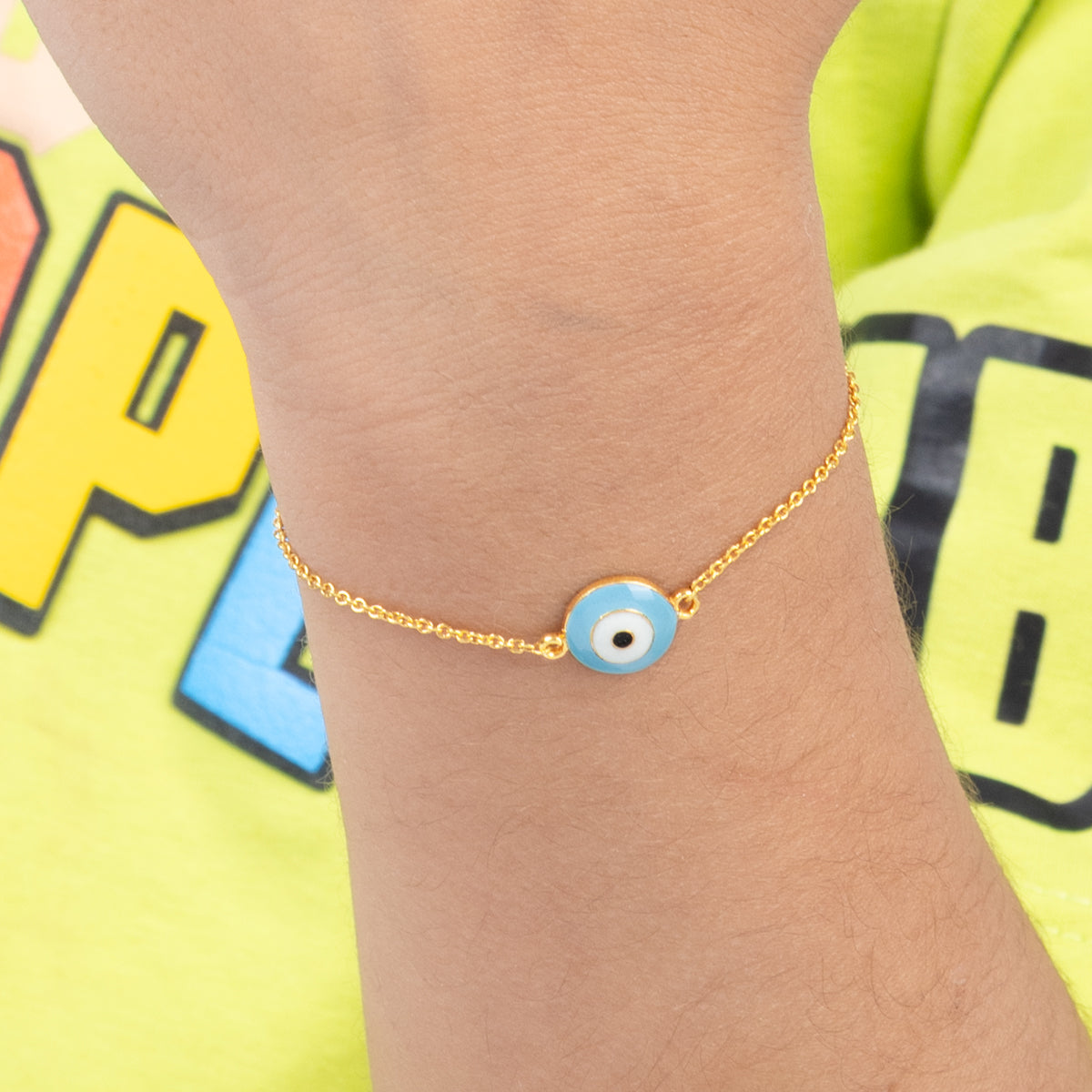 Tiny Jewels gold bracelet with a blue evil eye charm on a wrist, against a colorful background.