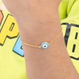 Tiny Jewels gold bracelet with a blue evil eye charm on a wrist, against a colorful background.