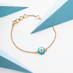 Tiny Jewels gold bracelet with a blue evil eye charm on a white and blue geometric background