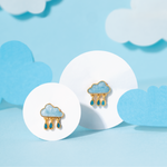 Tiny Jewels pair of cloud-shaped earrings with blue accents on a light blue background with cloud decorations.