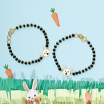 Tiny Jewels two black beaded bracelets with bunny charms on a blue background with grass and carrots.