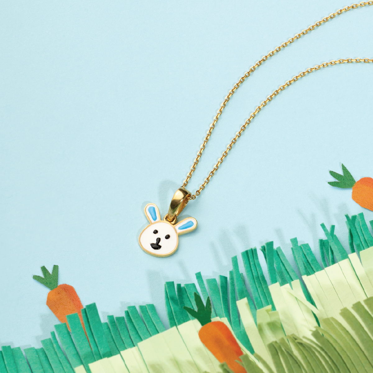 Tiny Jewels Gold necklace with a bunny pendant against a light blue background with cartoon carrots and grass.