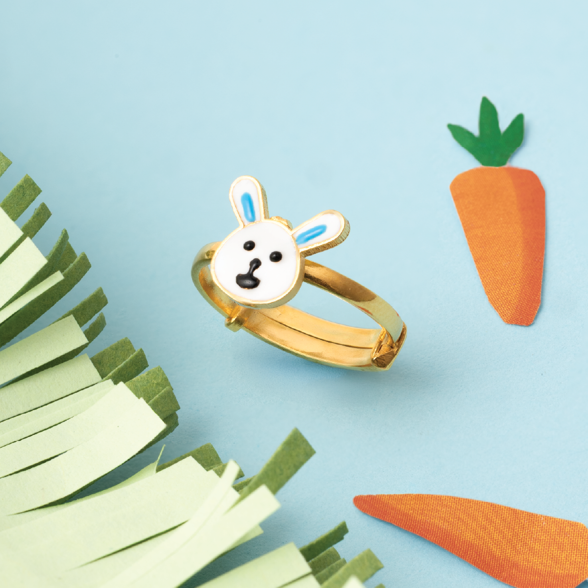 Tiny Jewels gold bunny ring with a white face on a blue background with paper carrots and grass.