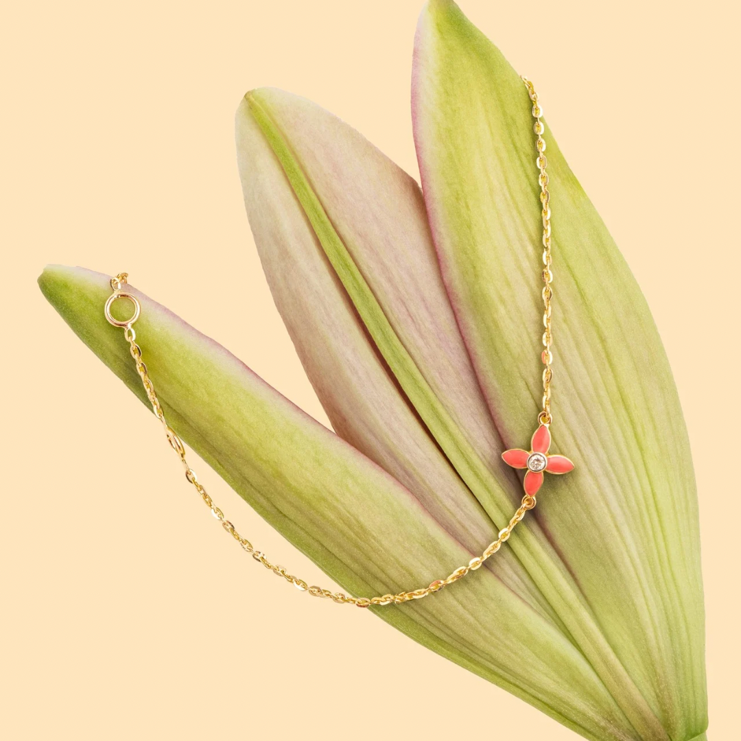 Tiny Jewels gold necklace with a red flower pendant on green leaves against a beige background