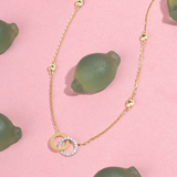 Tiny Jewels gold necklace with circular pendant on a pink background with green lemons