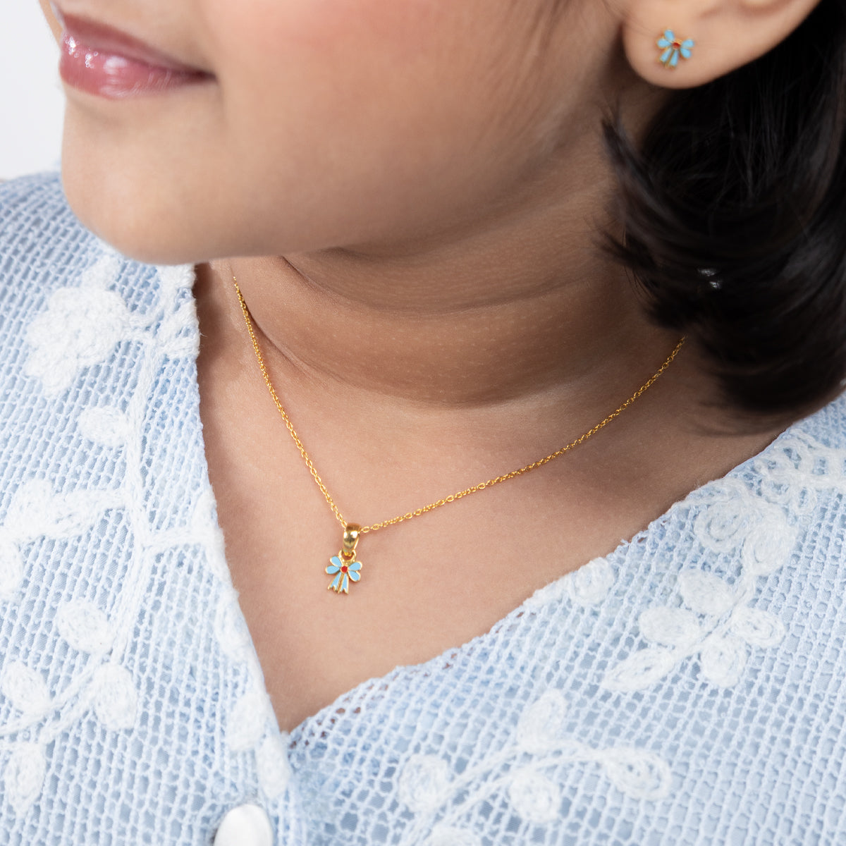 Tiny Jewels close-up of a person wearing a gold necklace with a small pendant, against a light blue embroidered garment.