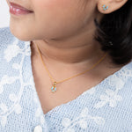 Tiny Jewels close-up of a person wearing a gold necklace with a small pendant, against a light blue embroidered garment.