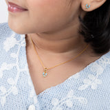 Tiny Jewels close-up of a person wearing a gold necklace with a small pendant, against a light blue embroidered garment.