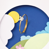 Tiny Jewels gold bracelet with a cartoon character on a colorful landscape background