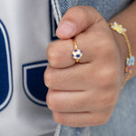 Tiny Jewels close-up of a hand wearing a gold ring with a blue and white design, against a blurred background.