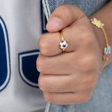 Tiny Jewels close-up of a hand wearing a gold ring with a blue and white design, against a blurred background.
