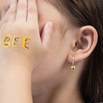 Tiny Jewels close-up of a person wearing a gold bee-shaped earring with a blurred background.