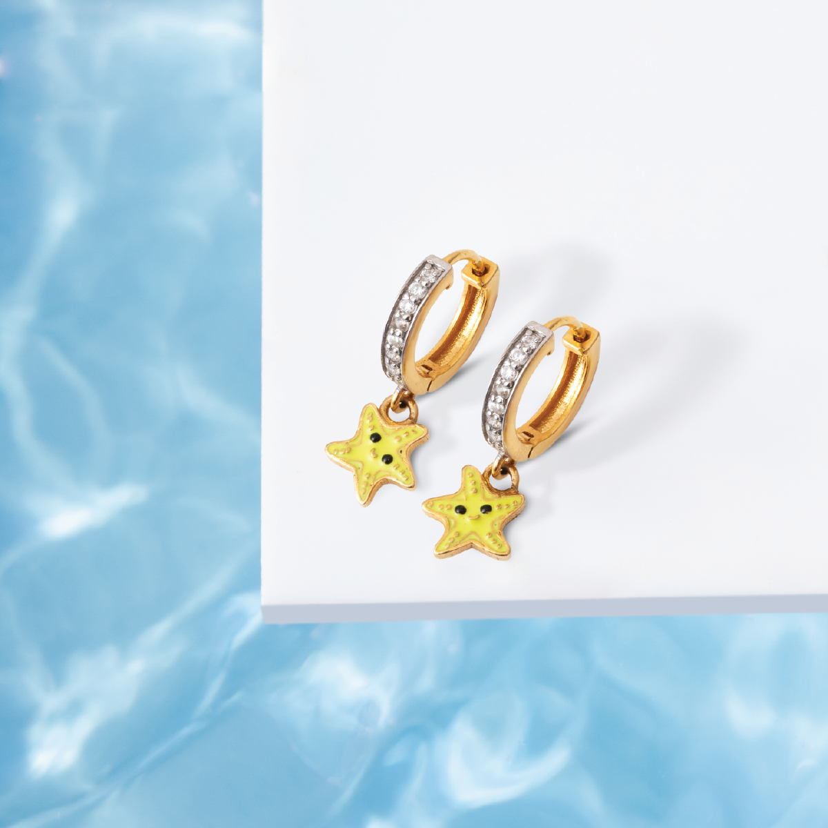 Tiny Jewels gold hoop earrings with star-shaped charms on a white card against a blue water background