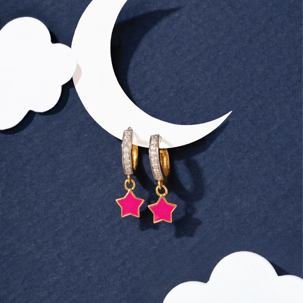 Tiny Jewels gold hoop earrings with pink star charms on a blue background with a white crescent moon and cloud.