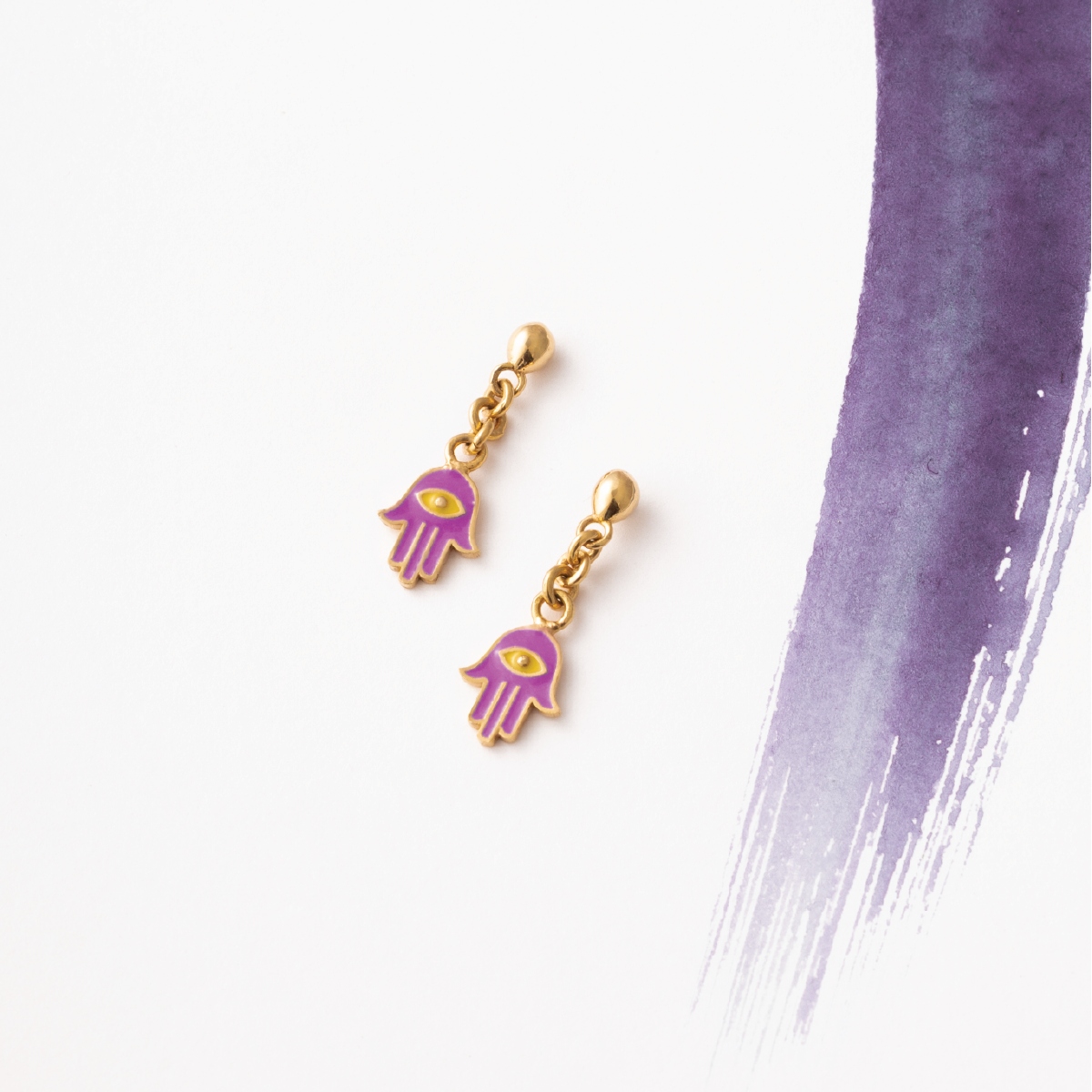 Tiny Jewels gold and purple hamsa hand earrings on a white background with a purple brushstroke.