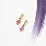 Tiny Jewels gold and purple hamsa hand earrings on a white background with a purple brushstroke.