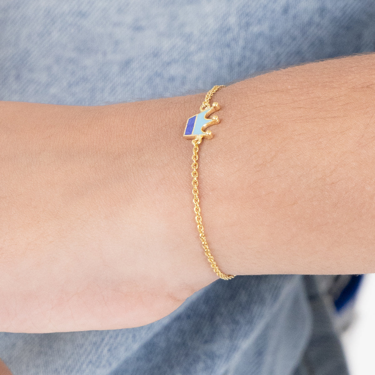 Tiny Jewels gold bracelet with a blue and white heart charm on a wrist against a denim background