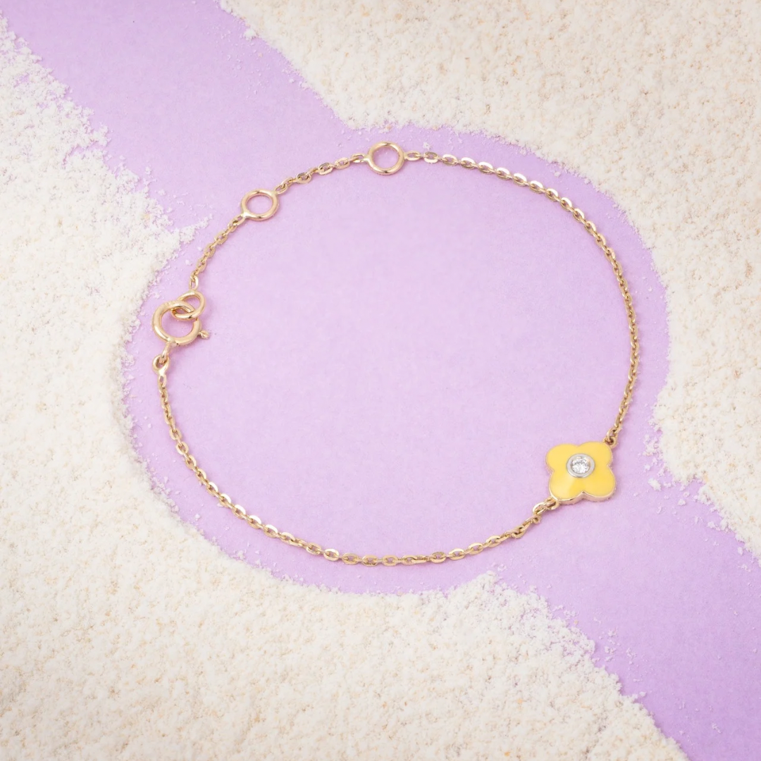 Tiny Jewels gold bracelet with a yellow flower charm on a textured purple and beige background