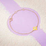 Tiny Jewels gold bracelet with a yellow flower charm on a textured purple and beige background