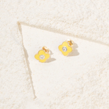Tiny Jewels yellow flower-shaped earrings with a clear gemstone on a textured beige surface