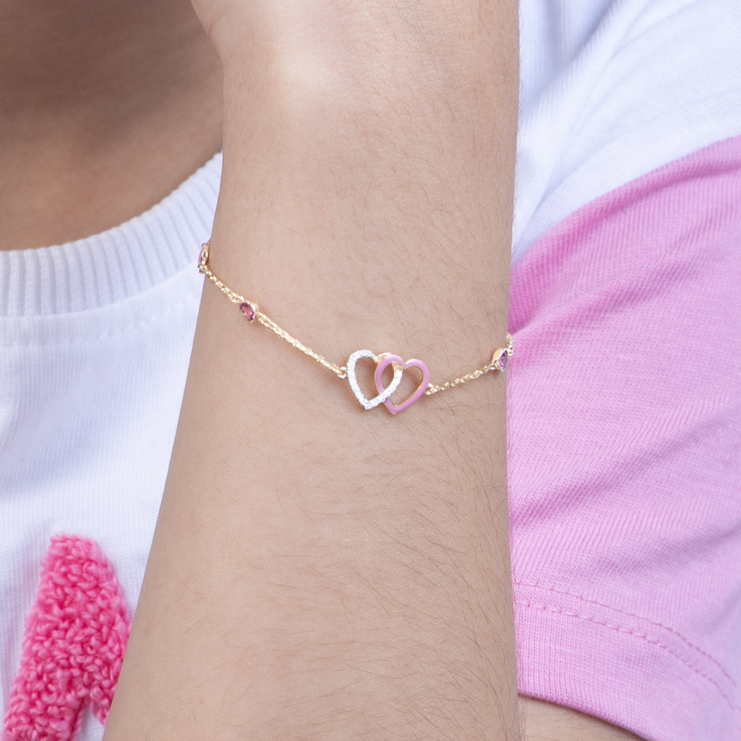 Tiny Jewels gold bracelet with interlocking heart charms on a wrist against a white and pink background