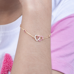 Tiny Jewels gold bracelet with interlocking heart charms on a wrist against a white and pink background