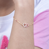 Tiny Jewels gold bracelet with interlocking heart charms on a wrist against a white and pink background