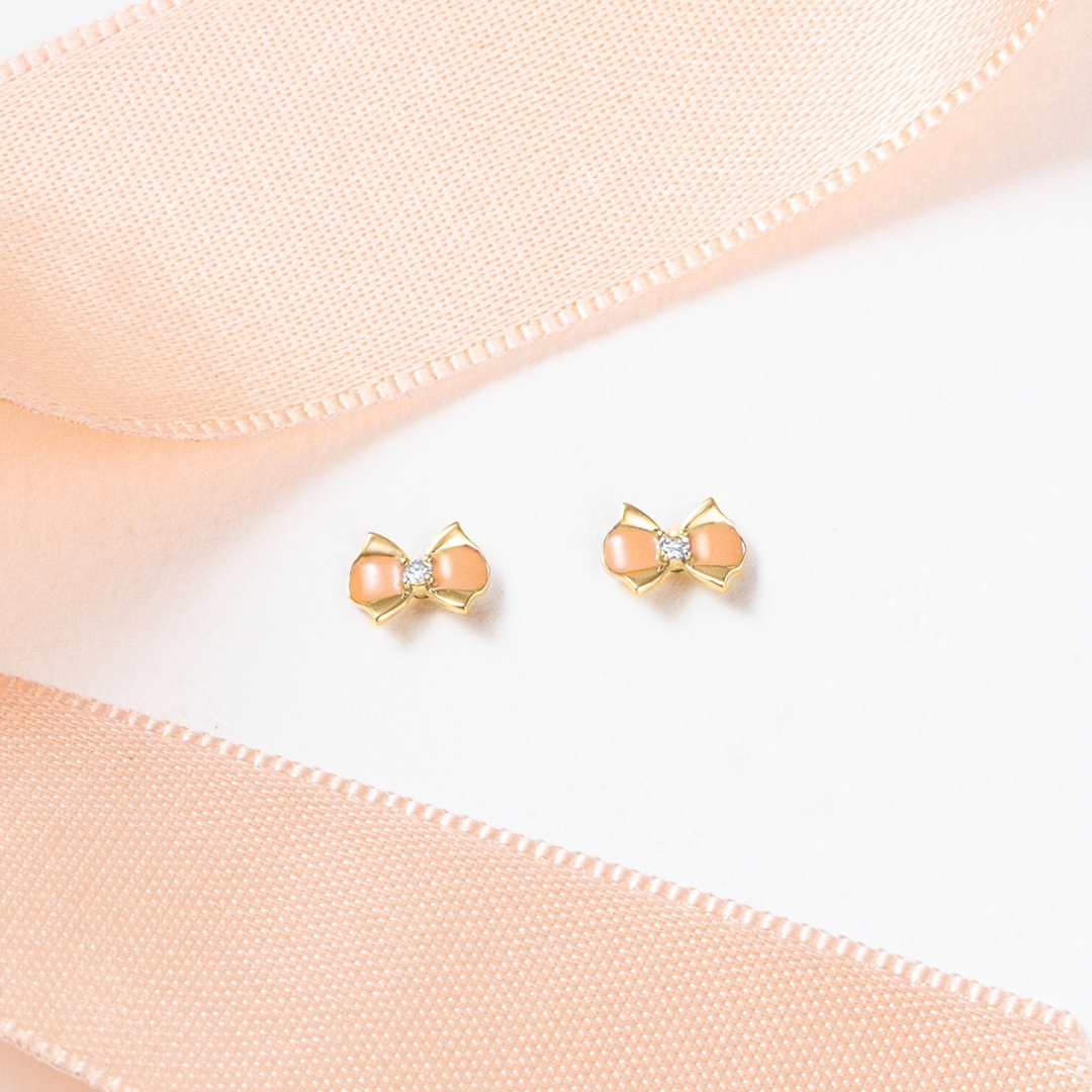 Tiny Jewels pair of bow-shaped earrings with pink centers on a light pink fabric background.