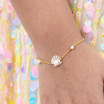 Tiny Jewels gold bracelet with a shell and pearl charm on a wrist against a colorful sequin background
