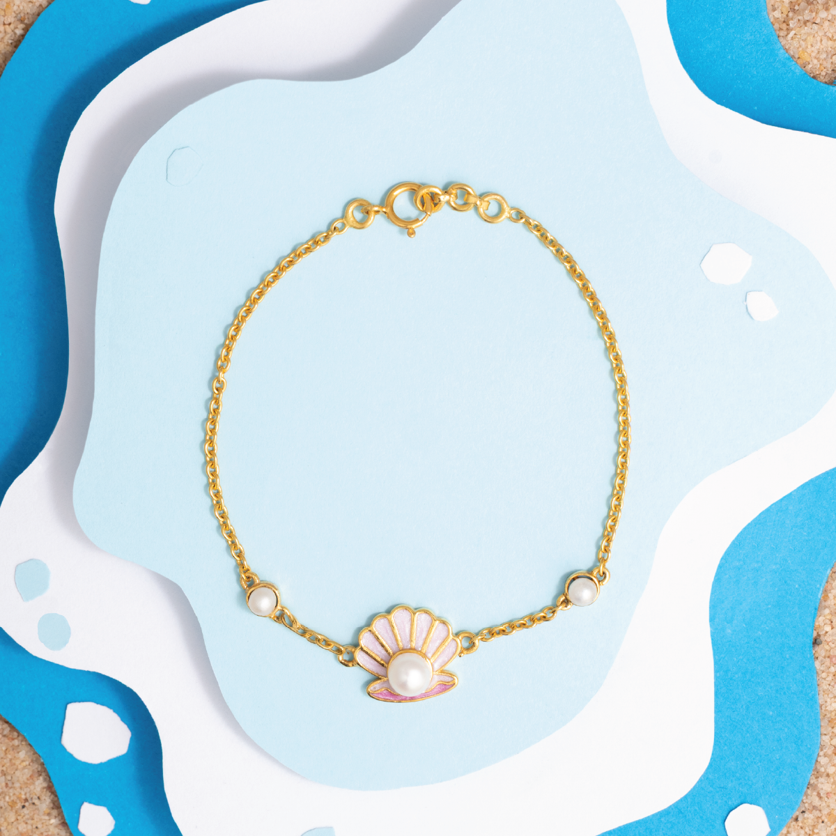 Tiny Jewels gold bracelet with a shell and pearl charm on a blue and white abstract background