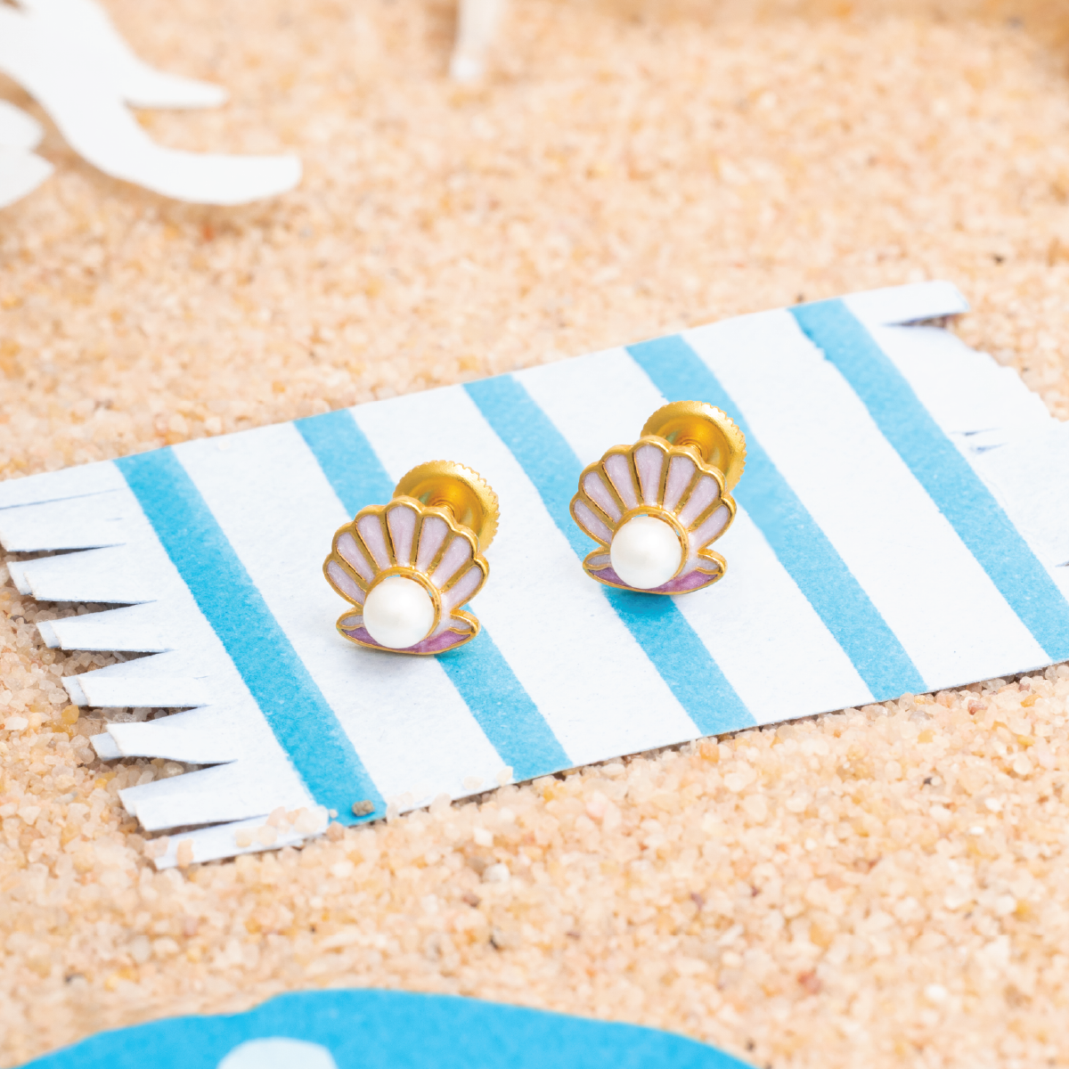 Tiny Jewels shell-shaped earrings on a striped card with sand background