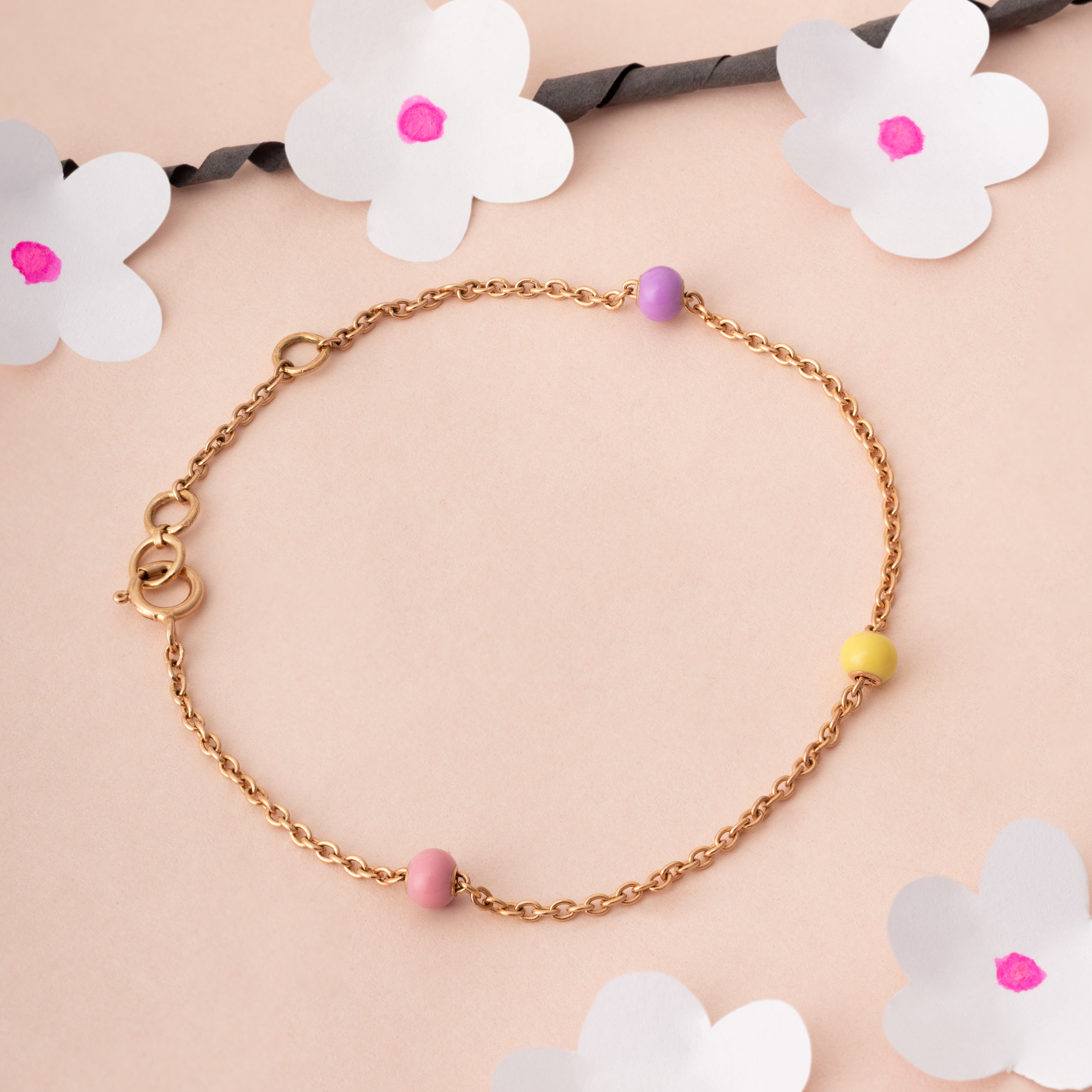 Tiny Jewels gold bracelet with colorful beads on a pink background with floral decorations