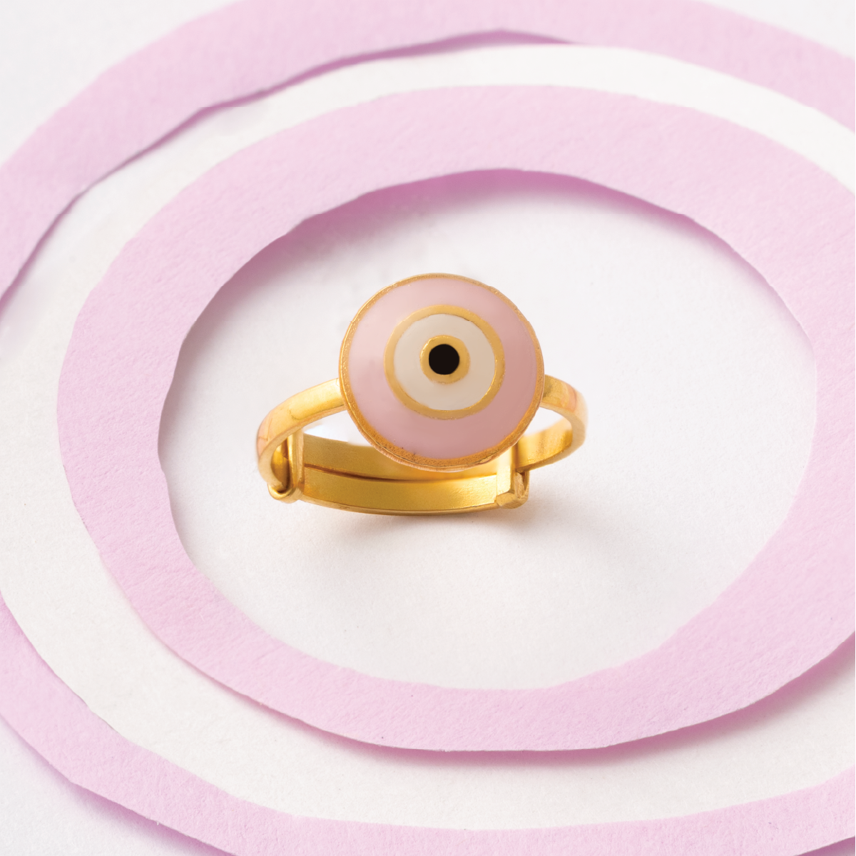 Tiny Jewels gold ring with a pink evil eye design on a pink and white circular background