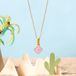 Tiny Jewels gold necklace with a pink flower pendant against a beach background with palm trees and cacti.