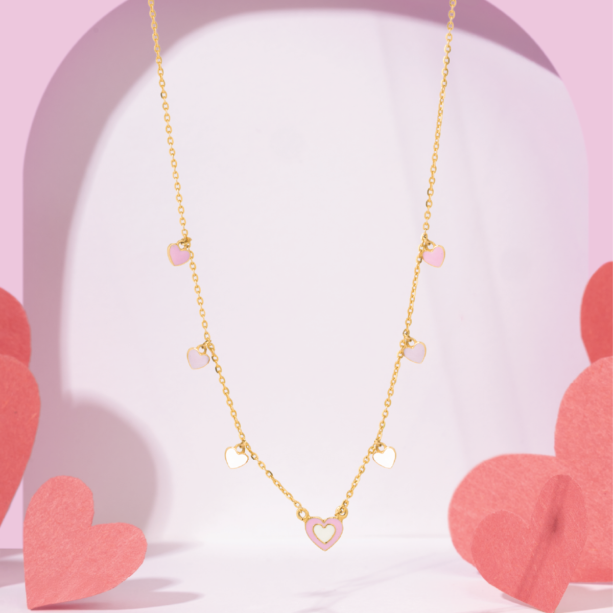 Tiny Jewels gold necklace with heart-shaped pendants on a pink heart-shaped background